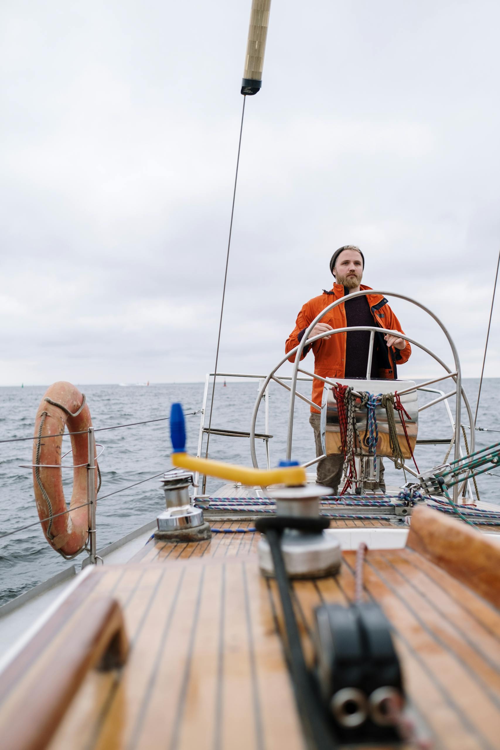 Man in orange jacket steering a sailboat on the open sea, showcasing adventure and boating lifestyle.