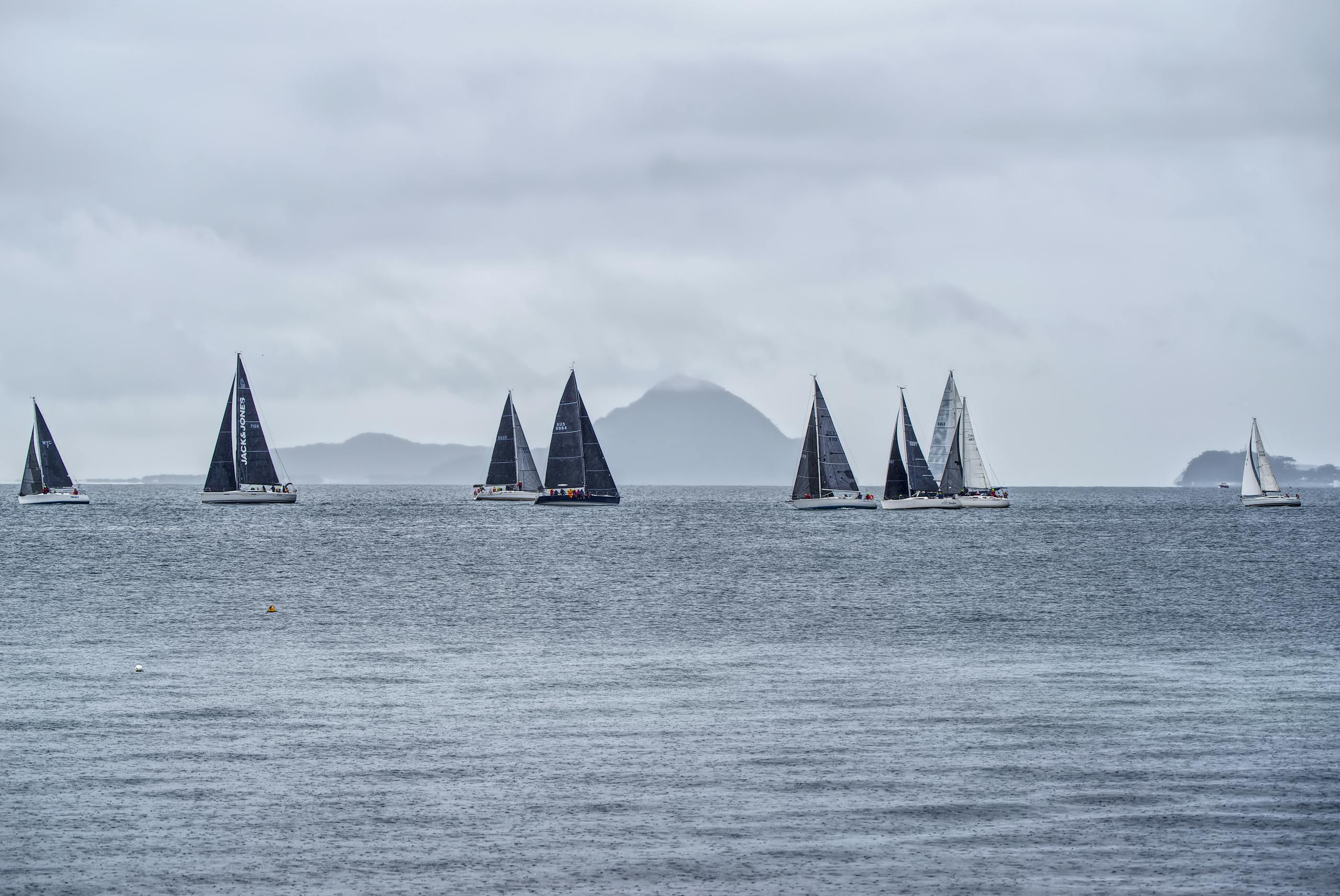 Multiple sailboats competing on the ocean with a mountain backdrop under a cloudy sky.